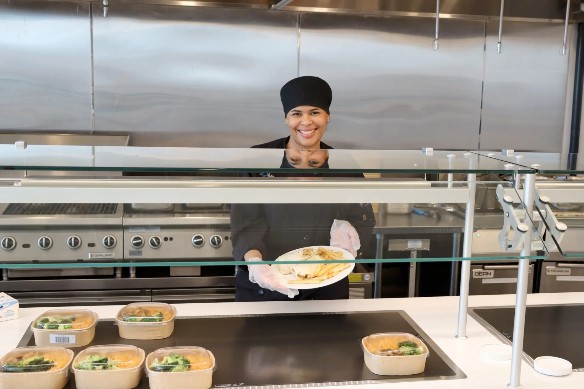 A smiling Columbia Dining employee stands behind the servery and displays a plate of food in her hands.