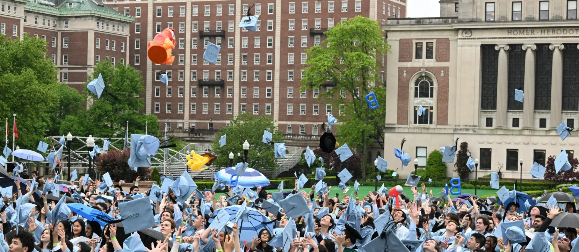 Graduating students in regalia throw their hats up in the air, with Butler Library, John Jay Hall, and Wallach Hall in the background.