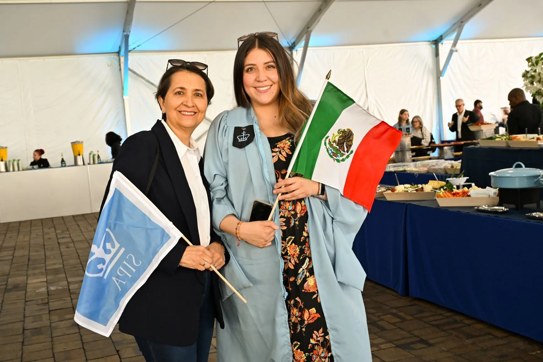A graduate and their family member stand inside a catering tent holding flags. 