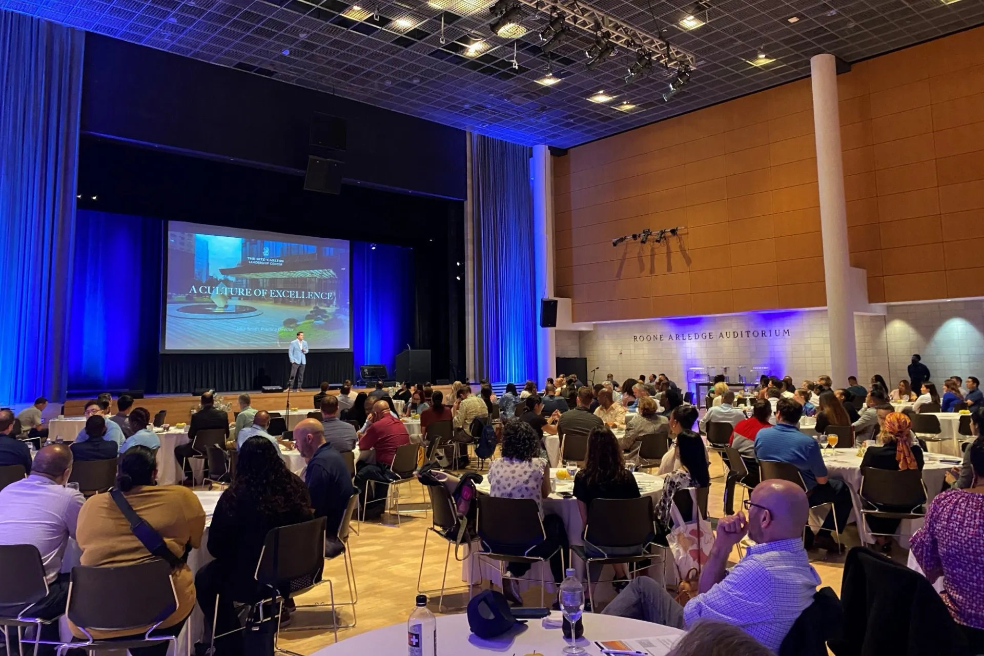A speaker addresses Facilities and Operations staff sitting at round tables in the Roone Arledge Auditorium of Lerner Hall.