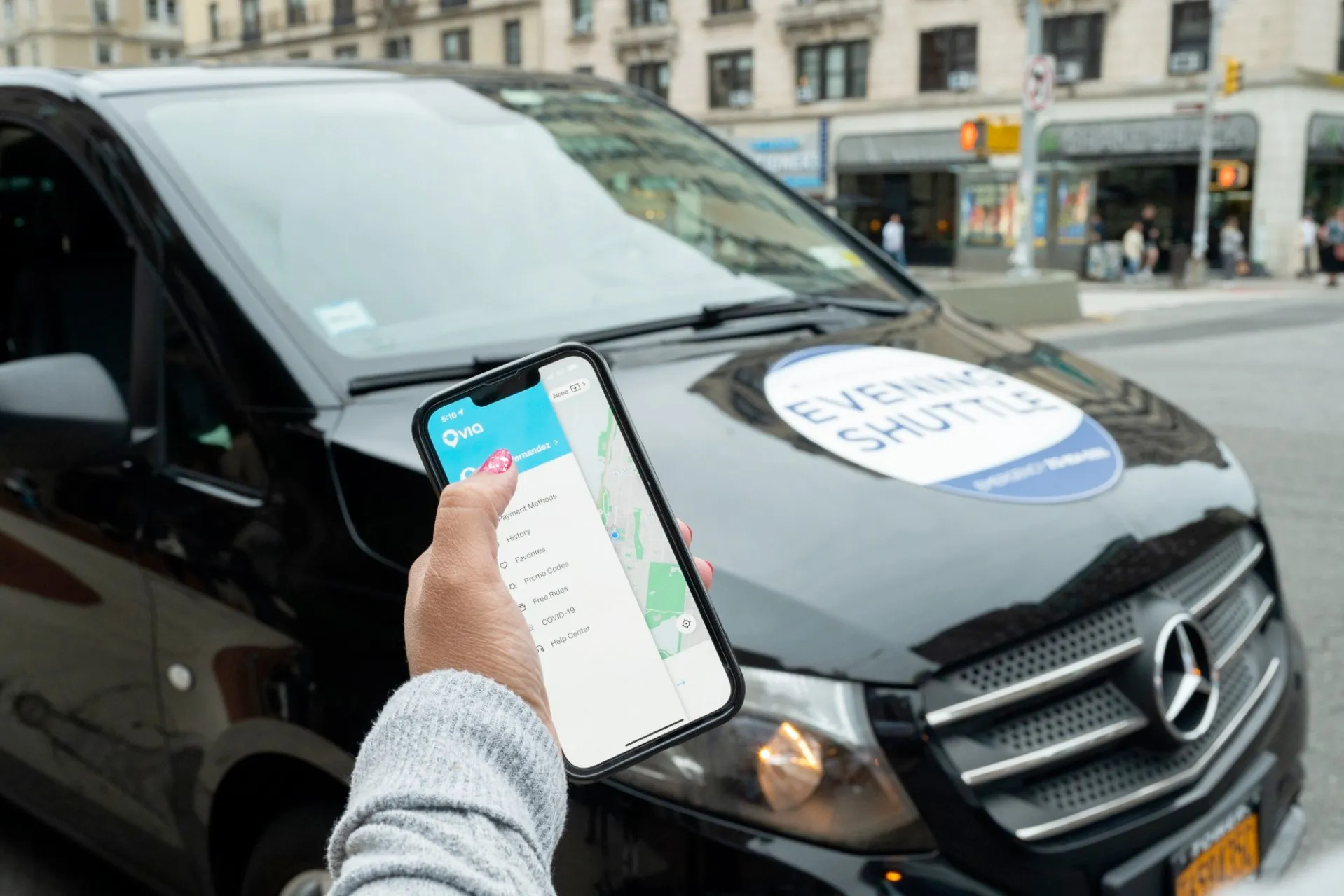 A person holds up a phone with the Via app loaded in front of a black van that has a big sticker on that says Evening Shuttle. 