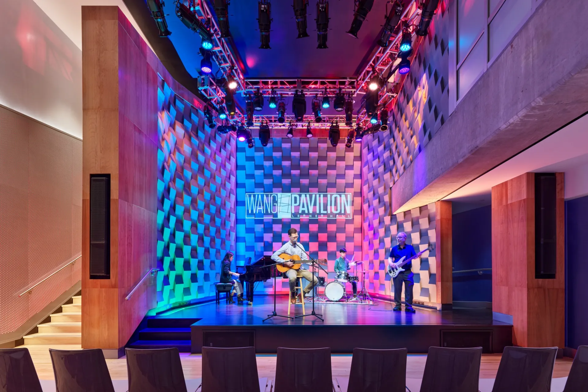 Wang Pavilion stage is lit up with blue, red, and purple lights. A band featuring a guitar player, pianist, and bass player is onstage performing.
